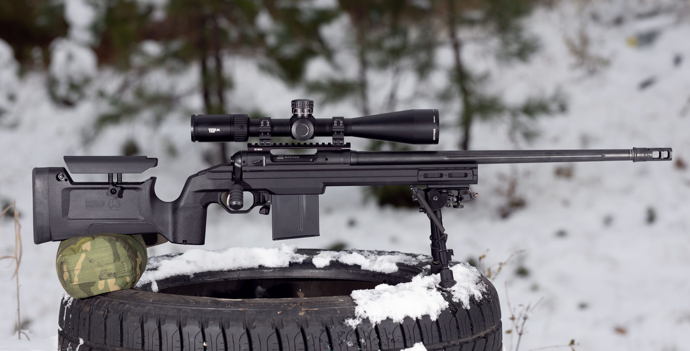 A black sniper rifle with a scope resting on a snow-covered tire outdoors.